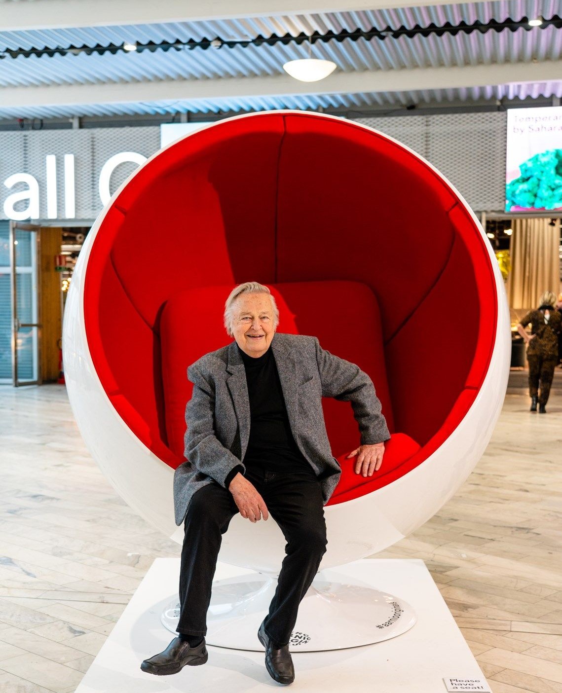Eero Aarnio assis dans sa Chaise Ball rouge et blanche. Il est habillé en noir avec une veste de costume grise. Cette chaise est en exposition dans un hall.